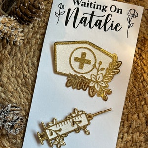 K&ouml;nnte beinhalten: Eine wei&szlig;e Karte mit dem Text "Waiting On Natalie" zeigt zwei goldfarbene medizinische Anstecknadeln. Eine Nadel ist eine Krankenschwesterm&uuml;tze mit einem Kreuz, die andere eine Spritze, beide mit floralen Designs verziert.