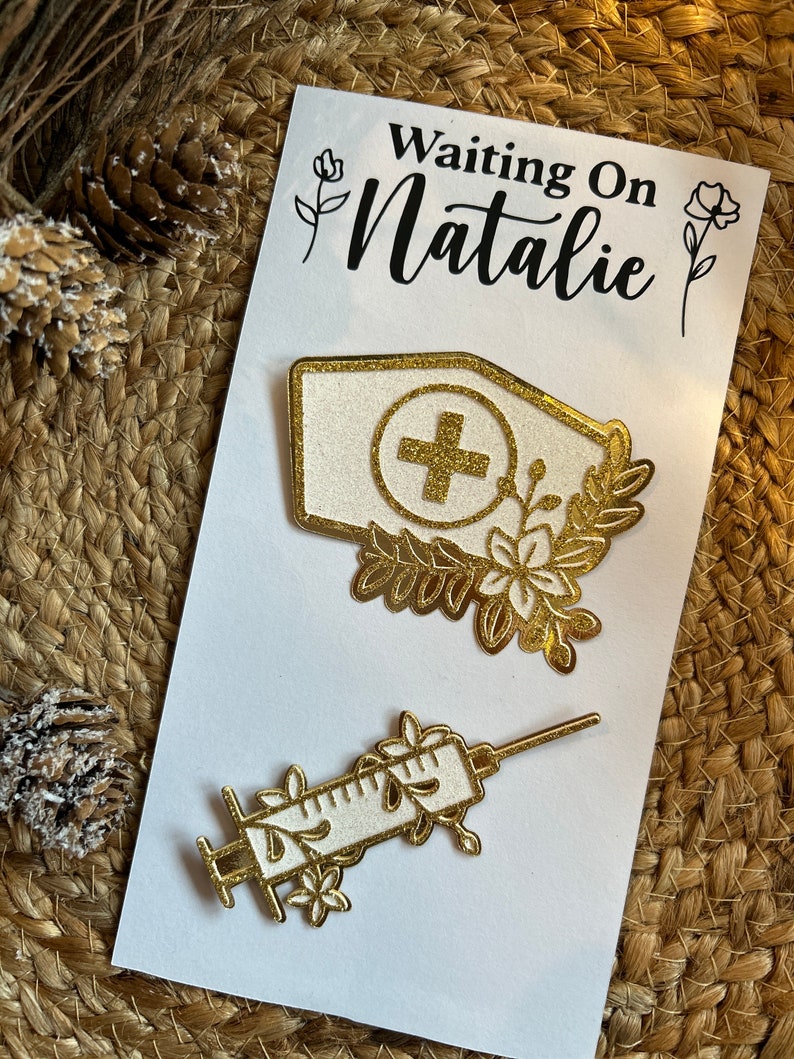 K&ouml;nnte beinhalten: Eine wei&szlig;e Karte mit dem Text "Waiting On Natalie" zeigt zwei goldfarbene medizinische Anstecknadeln. Eine Nadel ist eine Krankenschwesterm&uuml;tze mit einem Kreuz, die andere eine Spritze, beide mit floralen Designs verziert.
