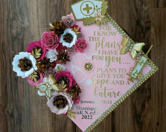 May include: Pink graduation cap decorated with paper flowers, gold accents, and a nurse's hat emblem. The cap features the text "For I know the plans I have for you..." and a stethoscope design. Includes the graduate's name and year.