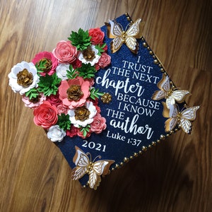 May include: A navy blue graduation cap decorated with paper flowers in shades of red, pink, white, and gold. Gold butterflies and the text "I trust the next chapter because I know the author Luke 1:37" are also featured.