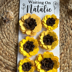 May include: A white card with the text "Waiting On Natalie" and six yellow and brown paper flowers. The flowers are arranged in two rows of three. The background is a woven, tan surface.