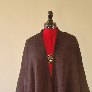 May include: A dark brown cape-style garment with a V-neck, displayed on a red dress form. The cape has a flowing design and appears to be made of a thick, textured fabric. The dress form is on a black metal stand.