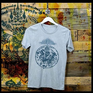 May include: A grey t-shirt with a black and white graphic print of a Thai Buddhist amulet. The amulet is surrounded by a circle of Thai script. The t-shirt is hanging on a wooden background.