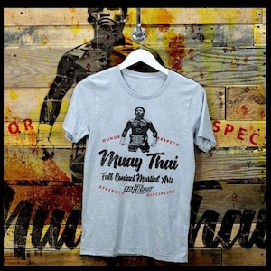 May include: A gray t-shirt with a vintage-style graphic of a Muay Thai fighter. The graphic includes the text "Muay Thai", "Full Contact Martial Arts", "Honor", "Respect", "Strength", and "Discipline".
