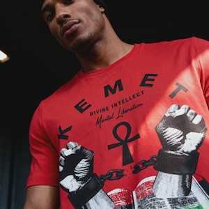 May include: Red t-shirt with the word "KEMET" in black block letters. The shirt features a graphic of two fists breaking chains, an ankh symbol, and the words "DIVINE INTELLECT Mental Liberation".