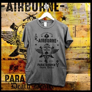 Puede incluir: Una camiseta gris con cuello redondo, colgada de una percha blanca. La camiseta presenta un gráfico negro con una calavera, alas y texto que dice "AIRBORNE", "DEALERS IN DEATH" y "PARATROOPER DEATH DEALER". El fondo es una pared de madera.