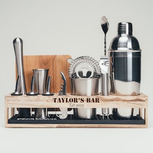 May include: A complete stainless steel bar tool set, including a shaker, strainer, and jigger, displayed in a wooden rack. The rack is engraved with "TAYLOR'S BAR Est 2023". The set also includes a cutting board, muddler, and bar spoon.
