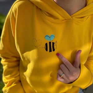 Könnte beinhalten: Ein gelber Hoodie mit einer schwarzen und gelben Biene mit blauen Flügeln, die auf der Vorderseite gestickt ist. Die Biene hält ein Herz in ihren Flügeln.