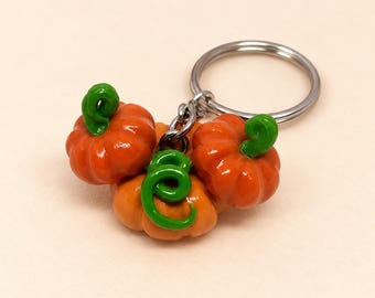May include: A keychain featuring three small, glossy orange pumpkins with bright green stems and curly vines attached to a silver metal split ring. The pumpkins are clustered together, showing detailed ridges and a smooth, reflective finish.