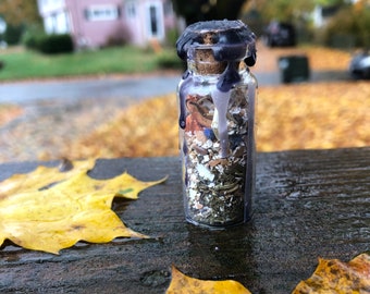 May include: A small glass bottle with a cork stopper is filled with dried herbs and flowers. The bottle is covered in a layer of dark purple wax, which has dripped down the sides. The bottle is sitting on a wooden surface with yellow leaves.