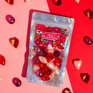 Pode incluir: Um saco de plástico transparente cheio de pérolas de banho vermelhas, cor-de-rosa e brancas. O saco tem um acabamento holográfico e um rótulo que diz "Retro Bath Pearls Pink Mix".