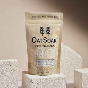Könnte beinhalten: Ein brauner Papierbeutel mit einem weißen Etikett, auf dem "Oat Soak Feed Your Skin" steht. Das Etikett zeigt auch eine Grafik mit zwei Weizenähren und eine Liste von Hauterkrankungen, für die das Produkt geeignet ist, darunter Trockenheit, Ekzeme, Empfindlichkeit und Psoriasis. Der Beutel liegt auf einer weißen, strukturierten Oberfläche.