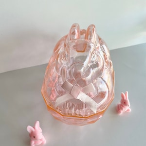 May include: A pink glass bunny-shaped trinket box with a matching bow. Two small pink bunny figurines are placed near the box. The box and figurines are on a white and gray surface.