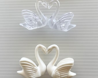 May include: Two pairs of swan-shaped decorative ornaments, one pair made of clear transparent material and one pair made of solid white material, positioned below a cluster of small white artificial flowers with thin stems.