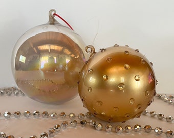 May include: Two glass Christmas ornaments, one iridescent gold and one gold with a textured surface, and a silver beaded garland.