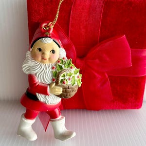 May include: A vintage Christmas ornament featuring a smiling gnome in a red outfit, white beard, and boots, holding a basket of berries. A red velvet gift box with a large bow is in the background. The ornament has a gold string for hanging.