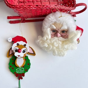 May include: Two Christmas ornaments, one is a wooden Rudolph the Red-Nosed Reindeer with a green wreath and a bell, the other is a Santa Claus head with a white beard and glasses. Both ornaments are hanging from a red wicker sleigh.