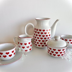 May include: A white tea set with a red heart pattern. The set includes a teapot, creamer, sugar bowl with lid, and two teacups and saucers.