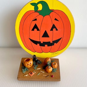 May include: A round wooden sign featuring a painted orange jack-o'-lantern on a yellow background with the text LYNN GATES, accompanied by a rectangular wooden base displaying miniature pumpkins, a black cat, and small candy-themed decorations.