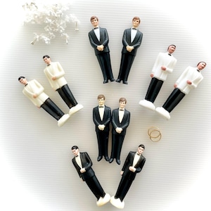 May include: A set of ten miniature groom figurines in black and white tuxedo attire. The figurines are arranged in a circle on a white surface. Two gold rings are in the center of the circle.