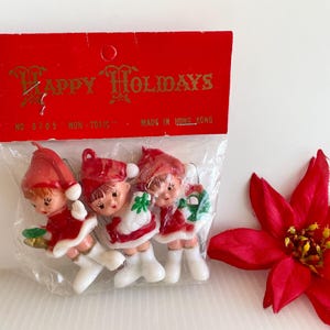 May include: A set of three plastic elf ornaments in a clear bag with a red header card labeled Happy Holidays. Each elf wears a red hat, red coat, and white boots. The header card includes the text No 8705, Non-Toxic, and Made in Hong Kong.