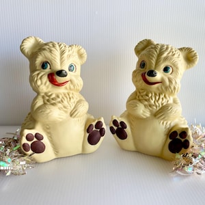 May include: Two vintage cream-colored bear figurines with blue eyes, red tongues, and brown paw pads. Each bear sits with its legs forward and has a fluffy chest. Silver tinsel accents the base of each bear. A charming, nostalgic collectible.