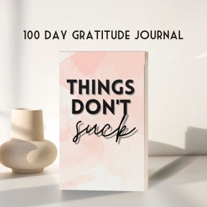 May include: A pink and white watercolor journal with the text "Things Don't Suck" in black. The journal is titled "100 Day Gratitude Journal".