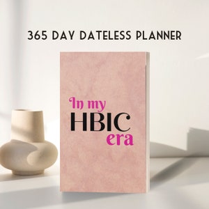 May include: A pink planner with the text "In my HBIC era" in black and pink lettering. The planner is a 365 day dateless planner.