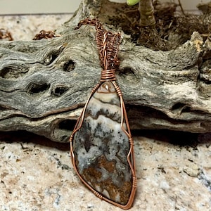 Pode incluir: Um pingente em forma de l&aacute;grima com uma pedra multicolorida estampada envolta em um fio de cobre decorativo. O pingente est&aacute; preso a uma corrente de cobre correspondente, exibindo detalhes intrincados de arame nas bordas da pedra.
