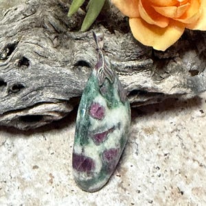 May include: A teardrop-shaped pendant featuring a polished stone with a mottled pattern of deep green, white, and vibrant magenta-pink inclusions. The stone is secured by a silver-toned metal bail at the top, showcasing a smooth, rounded surface.