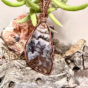 Pode incluir: Um pingente com uma pedra polida de formato irregular com padr&otilde;es manchados em marrom, branco e cinza escuro. A pedra &eacute; envolvida por um fio de cobre decorativo que forma uma argola no topo, prendendo a pedra dentro da estrutura met&aacute;lica.