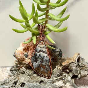 Pode incluir: Um pingente em forma de l&aacute;grima com uma pedra multicolorida de padr&otilde;es castanhos, brancos e pretos, envolta em fio de cobre decorativo. O pingente possui uma argola de fio torcido no topo para permitir a sua suspens&atilde;o.