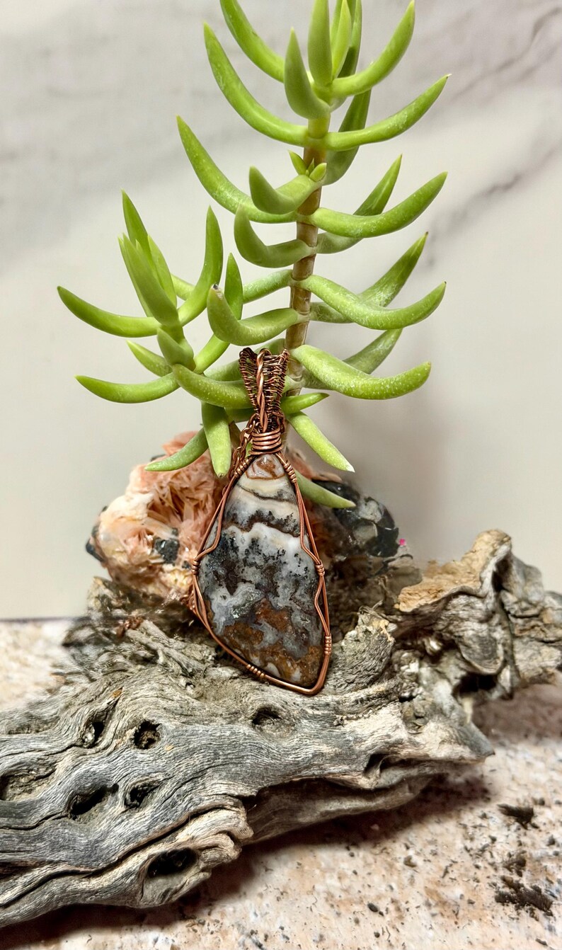 Pode incluir: Um pingente em forma de l&aacute;grima com uma pedra multicolorida de padr&otilde;es castanhos, brancos e pretos, envolta em fio de cobre decorativo. O pingente possui uma argola de fio torcido no topo para permitir a sua suspens&atilde;o.