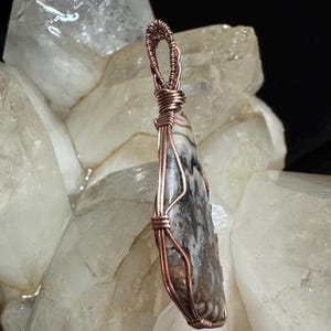 Pode incluir: Um pingente em forma de gota com uma pedra manchada em tons de castanho, branco e cinzento, envolvida por um fio de cobre decorativo. O fio forma uma moldura segura &agrave; volta da pedra e estende-se para cima numa pequena argola circular.