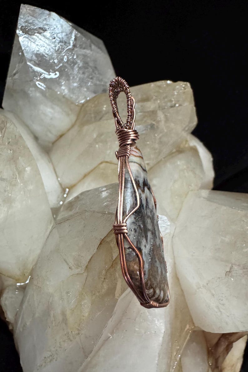 Pode incluir: Um pingente em forma de gota com uma pedra manchada em tons de castanho, branco e cinzento, envolvida por um fio de cobre decorativo. O fio forma uma moldura segura &agrave; volta da pedra e estende-se para cima numa pequena argola circular.