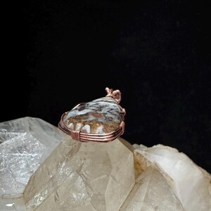 Pode incluir: Um pingente em forma de gota com uma pedra manchada em tons de castanho e branco, envolvida por um fio de cobre decorativo. O fio est&aacute; enrolado firmemente em torno do per&iacute;metro da pedra, formando um pequeno la&ccedil;o no topo para fixa&ccedil;&atilde;o.