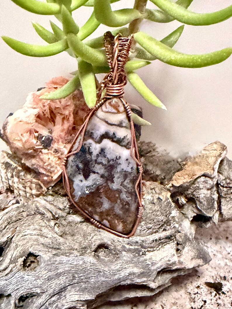 Pode incluir: Um pingente com uma pedra polida de formato irregular com padr&otilde;es manchados em marrom, branco e cinza escuro. A pedra &eacute; envolvida por um fio de cobre decorativo que forma uma argola no topo, prendendo a pedra dentro da estrutura met&aacute;lica.