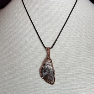 Pode incluir: Um colar com pendente apresentando uma pedra polida irregular com padr&otilde;es marmoreados em castanho, cinzento e branco. A pedra est&aacute; envolvida num fio de cobre decorativo e suspensa num cord&atilde;o preto simples, criando um contraste de texturas.