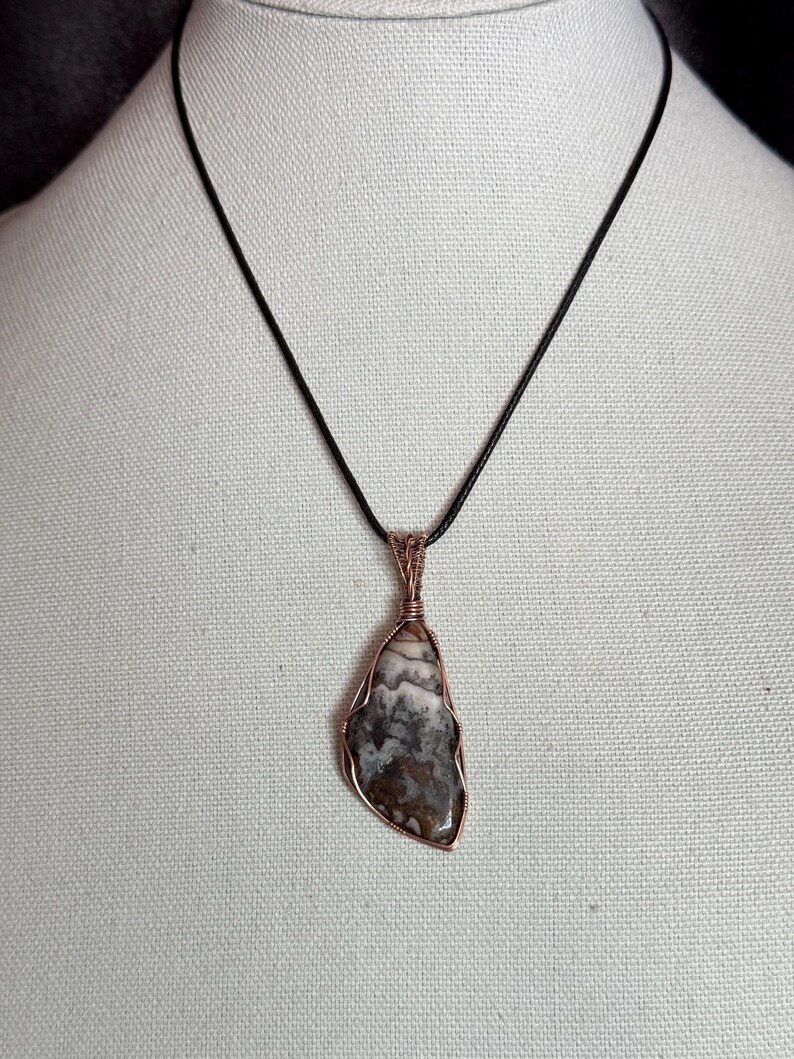 Pode incluir: Um colar com pendente apresentando uma pedra polida irregular com padr&otilde;es marmoreados em castanho, cinzento e branco. A pedra est&aacute; envolvida num fio de cobre decorativo e suspensa num cord&atilde;o preto simples, criando um contraste de texturas.