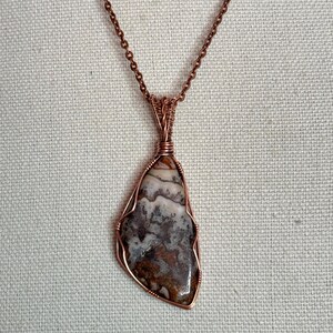 Pode incluir: Um colar com um pingente de pedra polida de formato irregular, apresentando padr&otilde;es em espiral nas cores castanho, branco e cinzento. A pedra est&aacute; presa numa arma&ccedil;&atilde;o de fio de cobre e suspensa por uma corrente de metal cor de cobre.