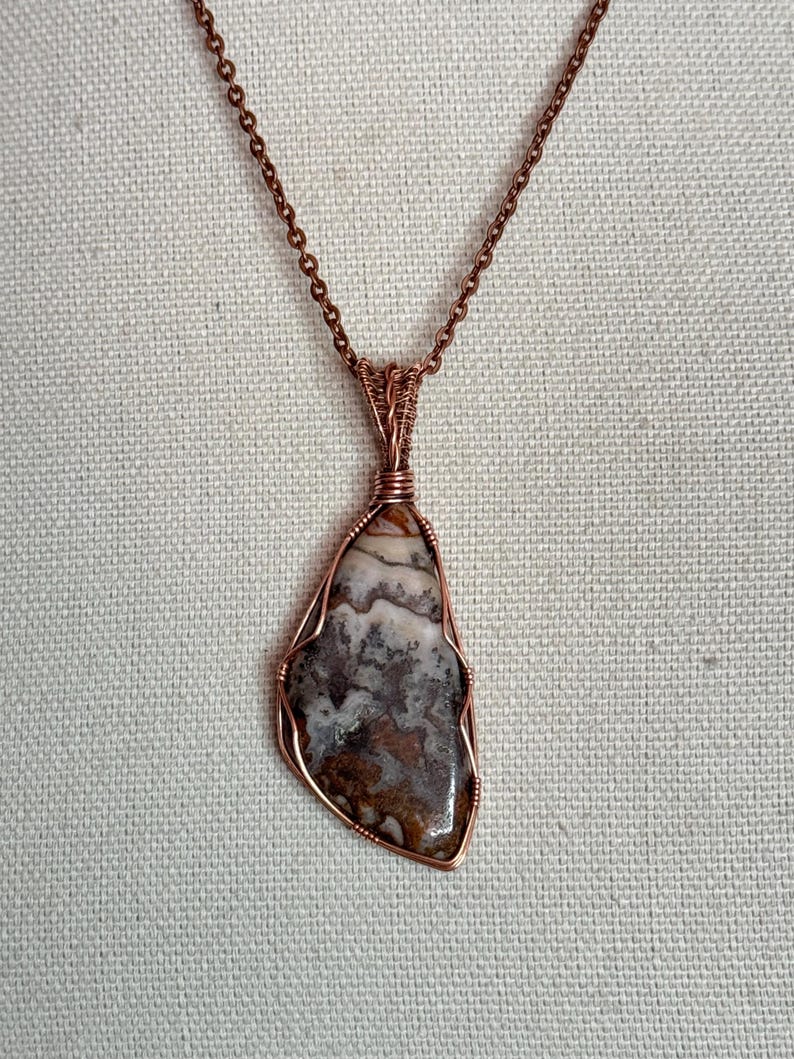 Pode incluir: Um colar com um pingente de pedra polida de formato irregular, apresentando padr&otilde;es em espiral nas cores castanho, branco e cinzento. A pedra est&aacute; presa numa arma&ccedil;&atilde;o de fio de cobre e suspensa por uma corrente de metal cor de cobre.