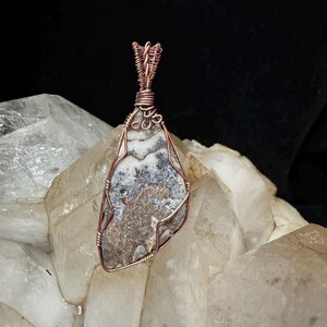 Pode incluir: Um pingente com uma pedra plana e irregular com padr&otilde;es manchados em cinza, branco e marrom, envolta em um fio de cobre decorativo. O fio forma uma moldura segura ao redor da pedra e se estende para cima em uma al&ccedil;a torcida para pendurar.