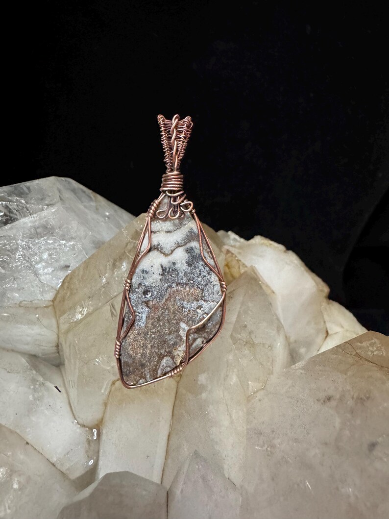 Pode incluir: Um pingente com uma pedra plana e irregular com padr&otilde;es manchados em cinza, branco e marrom, envolta em um fio de cobre decorativo. O fio forma uma moldura segura ao redor da pedra e se estende para cima em uma al&ccedil;a torcida para pendurar.