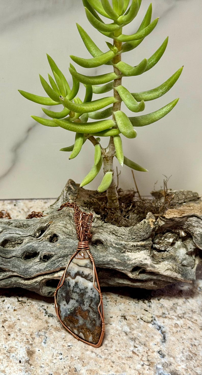 Pode incluir: Um pingente em forma de l&aacute;grima com uma pedra multicolorida estampada envolta em um fio de cobre decorativo. O pingente est&aacute; preso a uma corrente de cobre correspondente, exibindo detalhes intrincados de arame nas bordas da pedra.