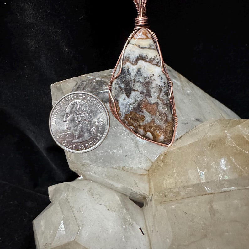 Pode incluir: Um pingente envolto em arame com uma pedra triangular multicolorida com padr&otilde;es de redemoinho marrons, brancos e cinzas. O fio de cobre envolve a pedra e estende-se para cima. Um quarto de d&oacute;lar americano est&aacute; colocado ao lado.