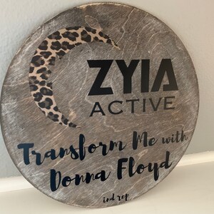 May include: A round wooden sign with a black leopard print crescent moon design. The sign says "ZYIA ACTIVE Transform Me with Donna Floyd ind rep".