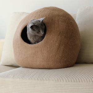 May include: A dome-shaped, tan felted cat cave with a circular opening. A grey cat is positioned inside the structure, looking out through the front entrance. The exterior surface of the pet bed has a soft, textured appearance and a rounded form.
