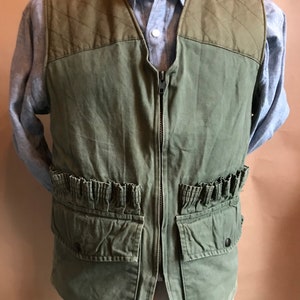 Könnte beinhalten: Eine grüne gesteppte Jagdjacke mit Reißverschluss und mehreren Taschen. Die Jacke hat einen Rüschenbesatz am unteren Rand und ein gestepptes Panel oben.