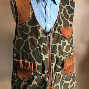 Könnte beinhalten: Eine Tarnjacke mit braunen Ledereinsätzen. Die Jacke hat einen Reißverschluss, zwei Taschen mit Klappen und zwei Patronenschlaufen an den Seiten.