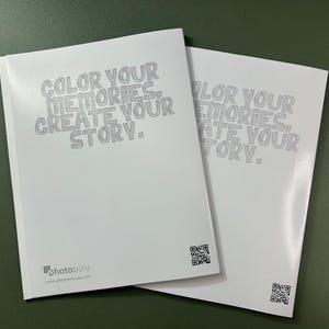 Może przedstawiać: Dwa puste białe albumy fotograficzne z tekstem "COLOR YOUR MEMORIES, CREATE YOUR STORY." nadrukowanym na okładce. Tekst jest w stylizowanym kroju pisma. Albumy mają kod QR na tylnej stronie.